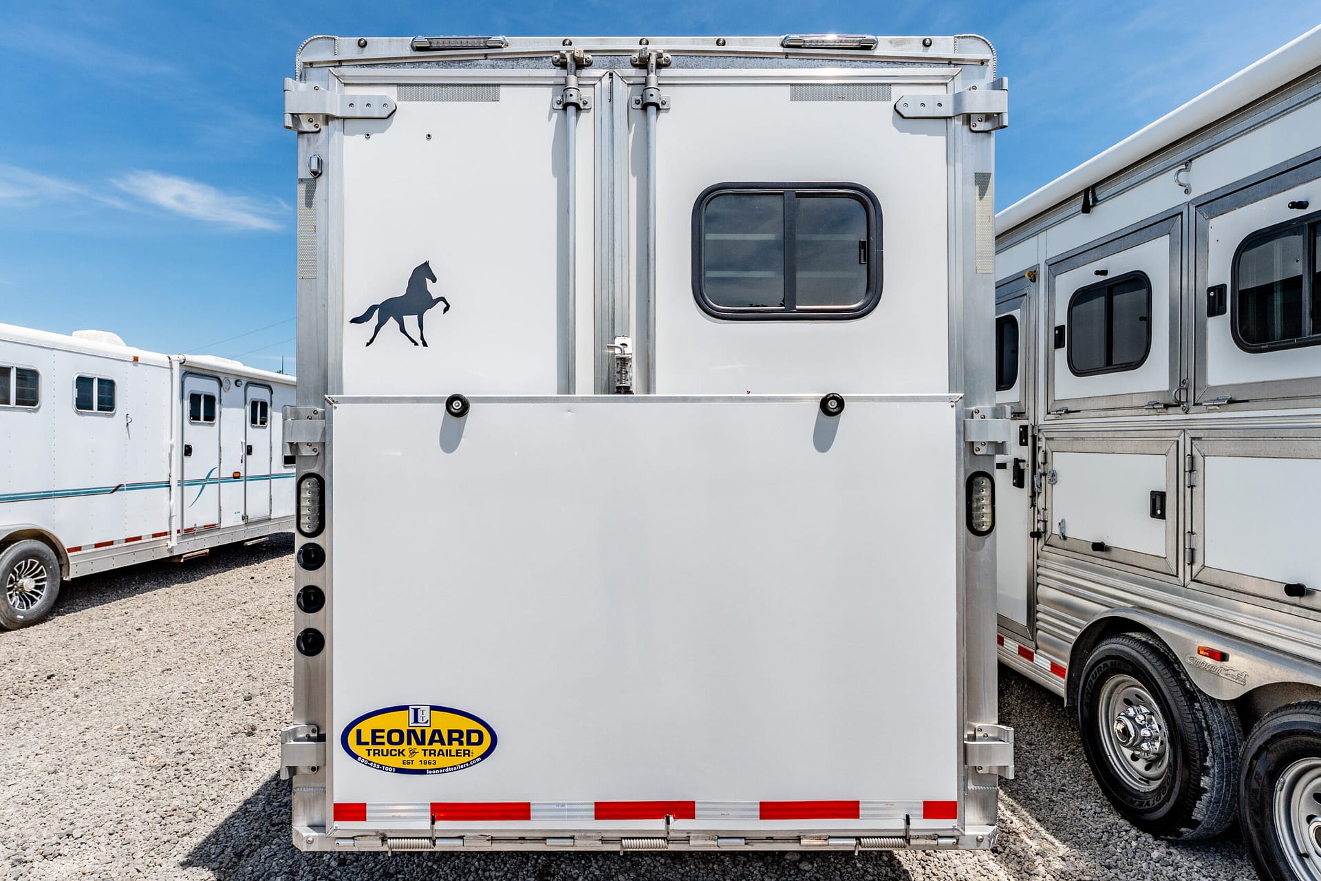 Rear view of a silver horse trailer with a black horse silhouette decal and a Leonard Truck & Trailer logo on the back edge.