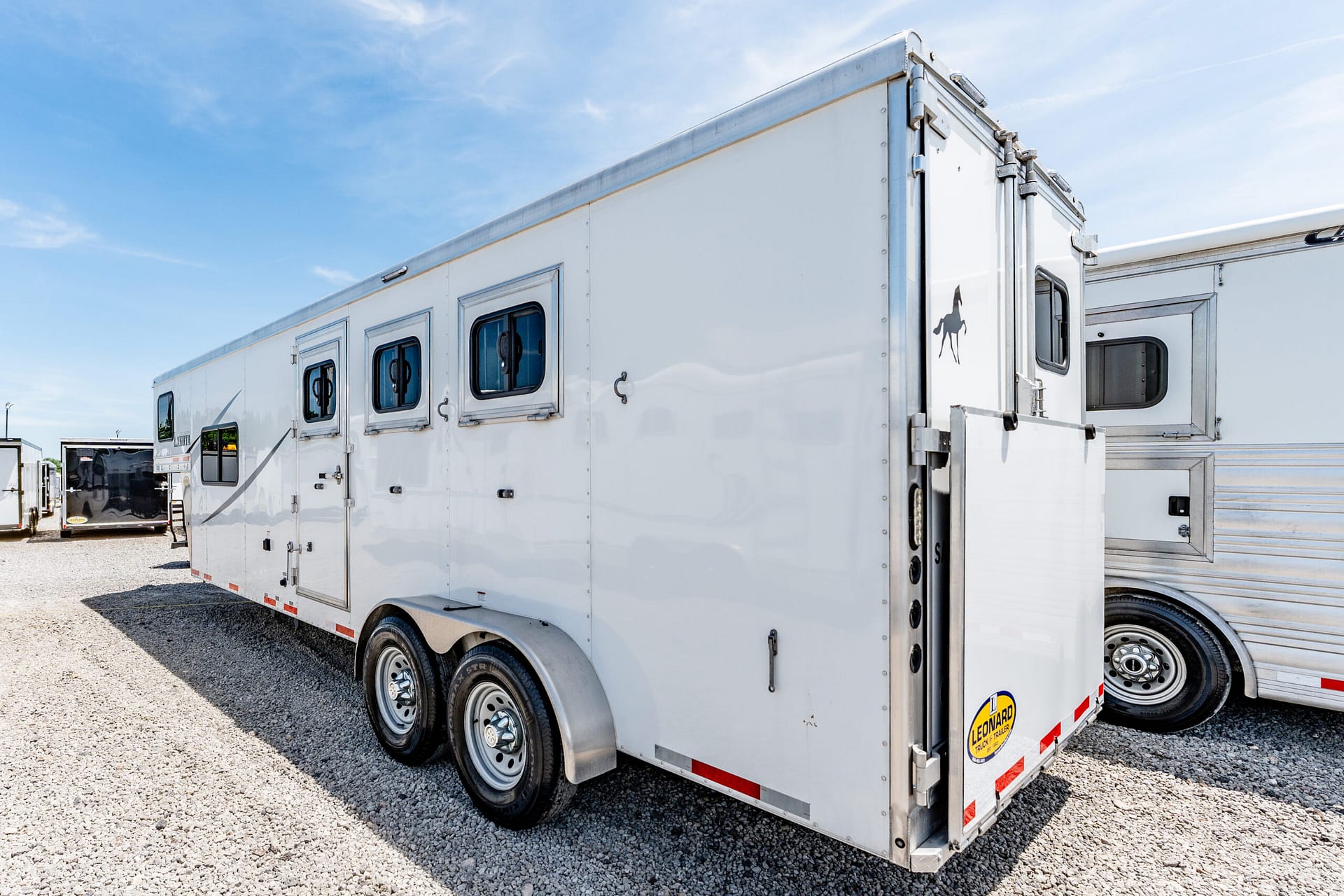A white semi-trailer parked in a gravel lot under a blue sky, with another trailer in the background and a horse logo on the rear door.
