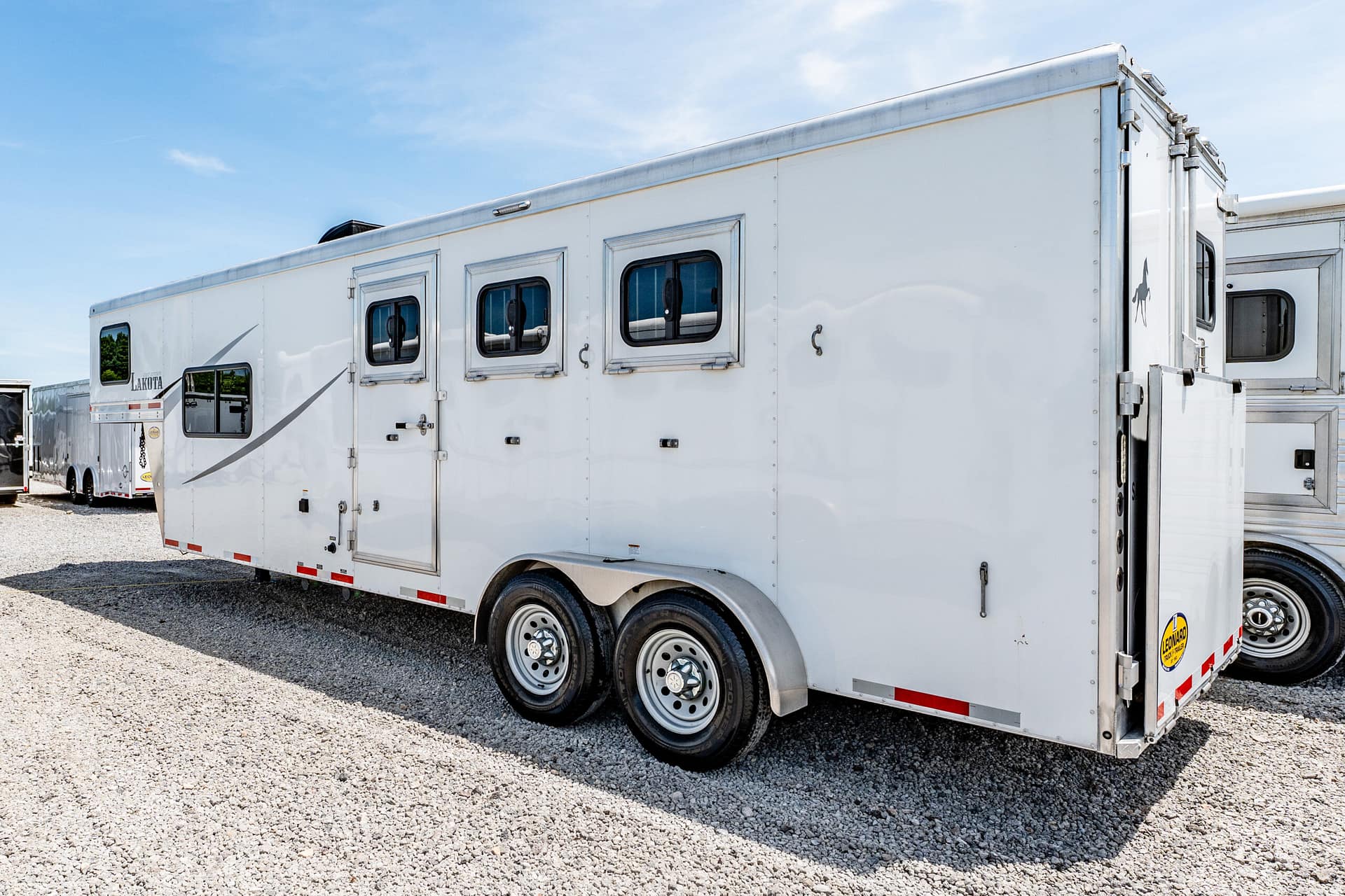 Long white horse trailer with multiple windows and dual axles parked on a gravel lot under a blue sky.