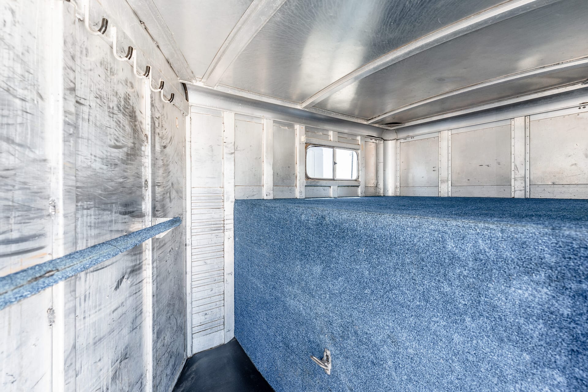 Interior of an empty aluminum trailer with blue carpeted floor and metal walls; hooks along left side and a small window at the far end.