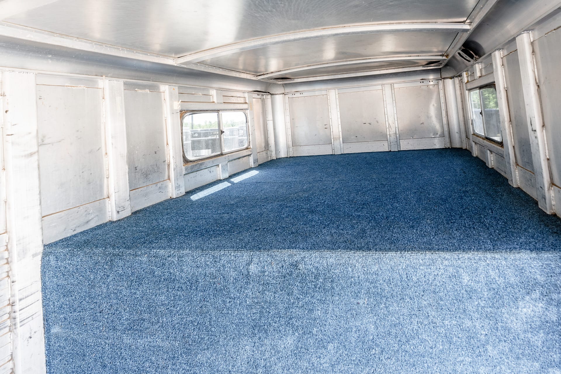 Empty metal-walled bus interior with blue carpet and windows along the sides.