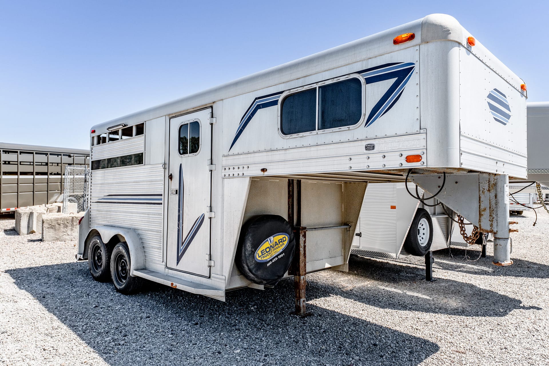 White horse trailer with blue graphics parked on a gravel lot under a clear blue sky.