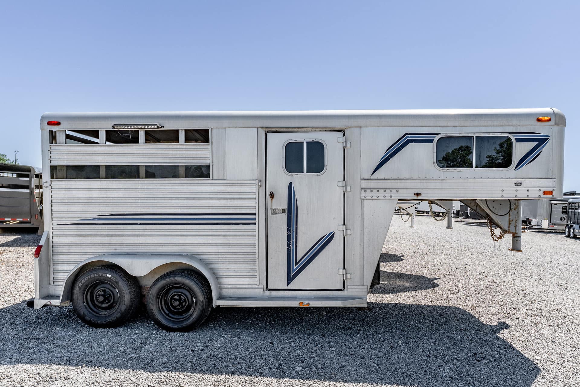 White horse trailer with blue stripes, parked on a gravel lot under a clear blue sky.