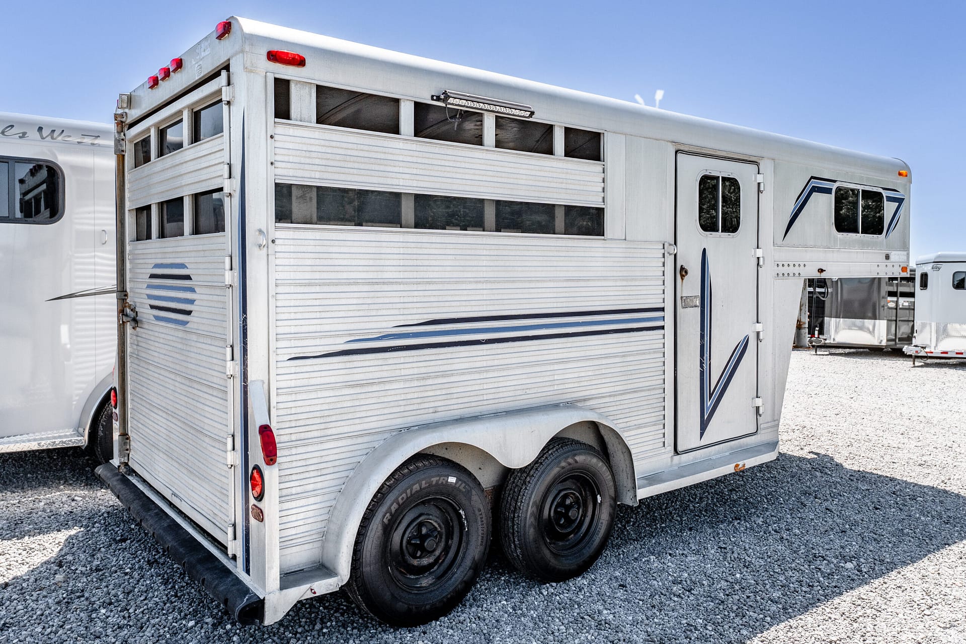 Silver aluminum horse trailer parked on a gravel lot under a clear blue sky, showing multiple side windows and dual rear wheels on the left side edge of the frame.