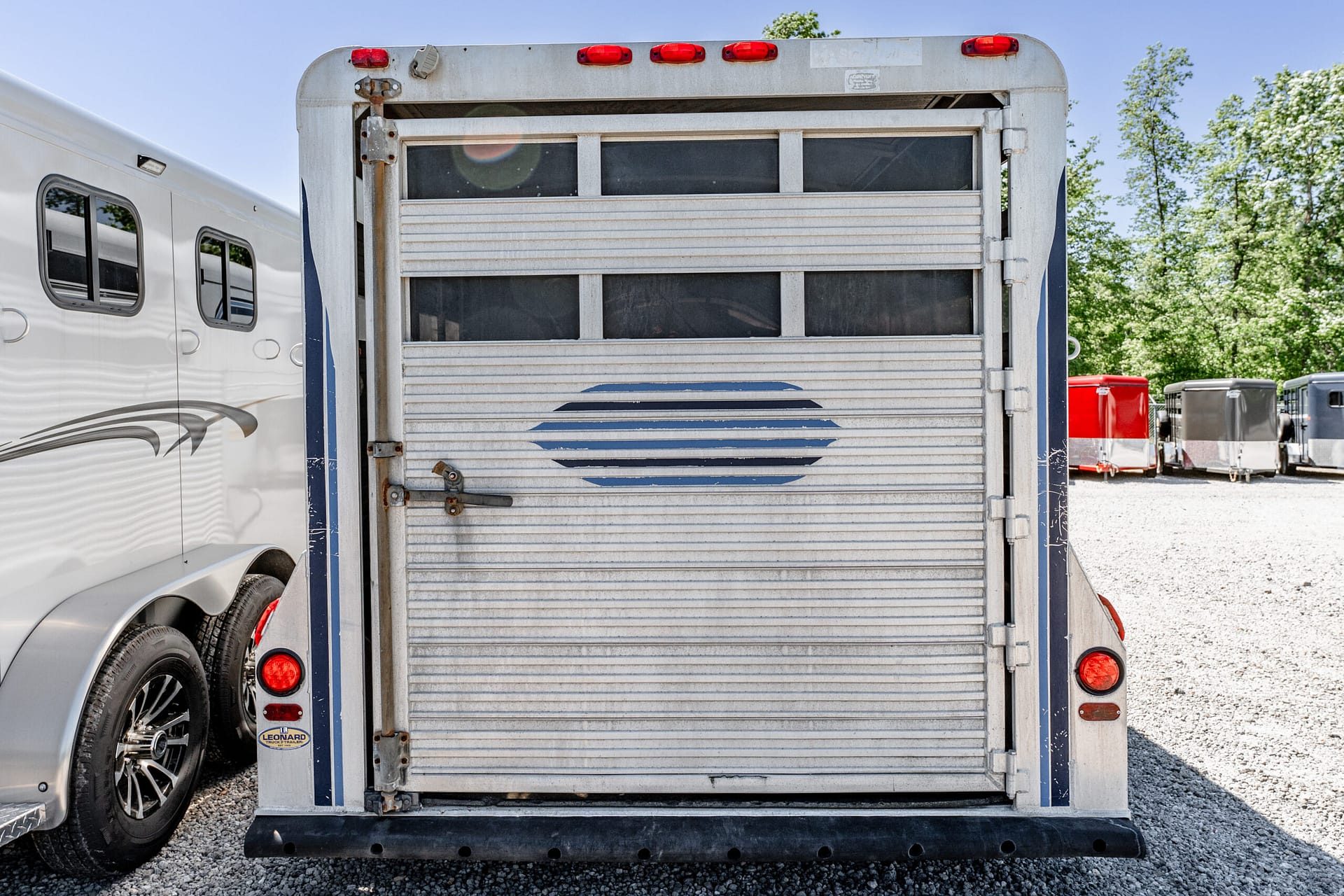 Rear view of a weathered metal trailer with a closed gate, red taillights, and a row of other trailers in a gravel lot.