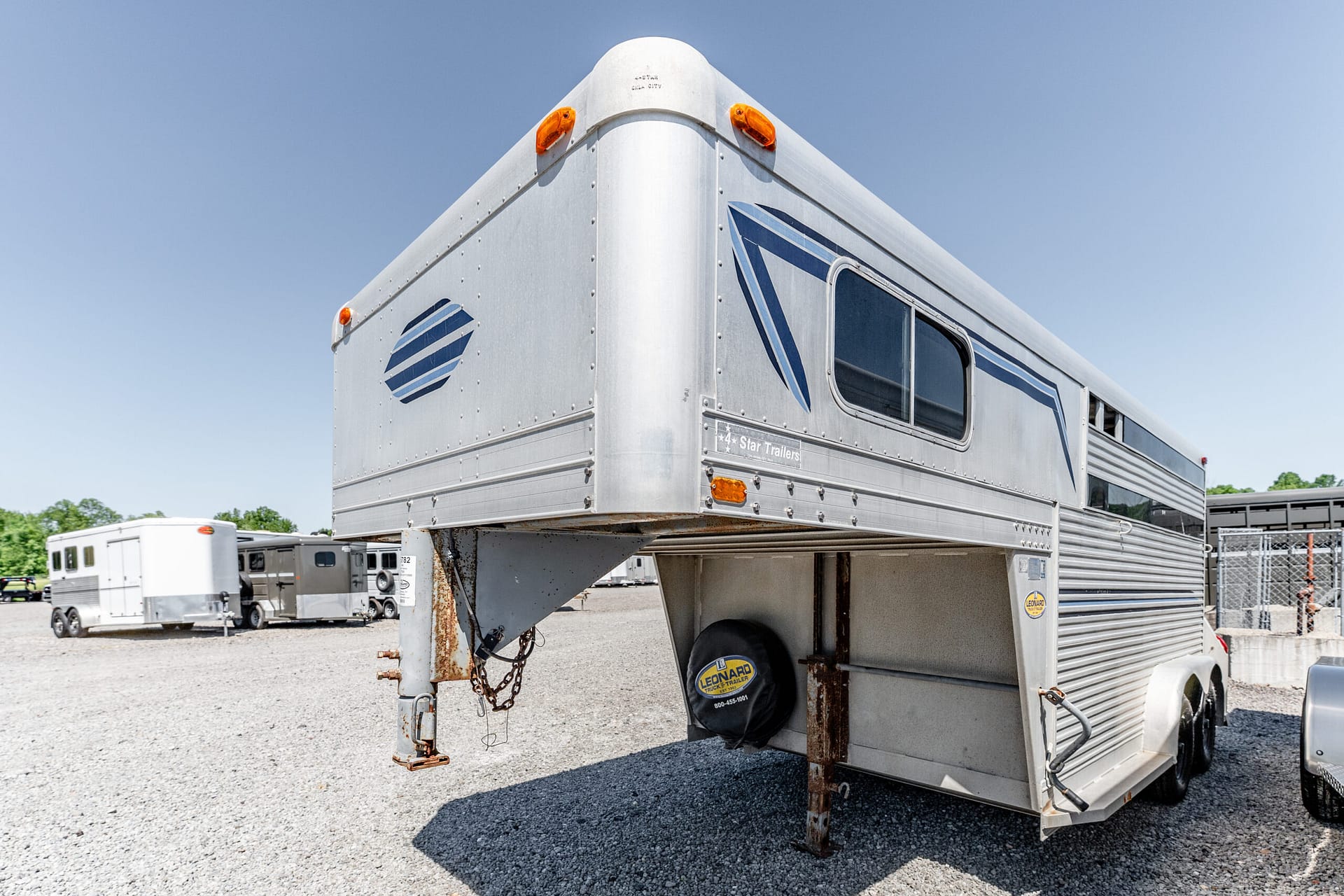 Silver gooseneck horse trailer with orange marker lights on a gravel lot, blue sky above