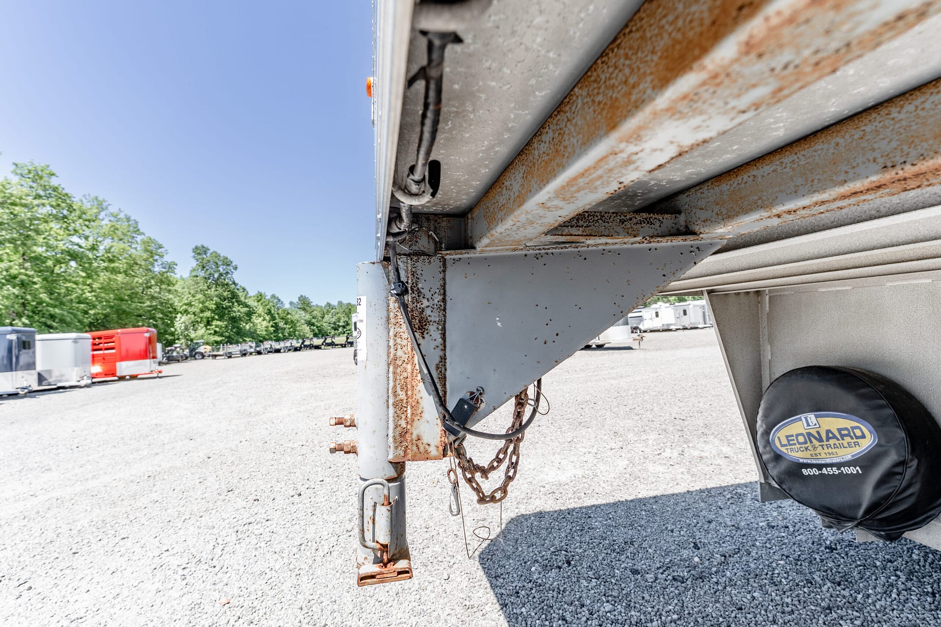 Close-up of a rusted trailer jack and metal frame with hanging chains on a gravel lot under sunny sky.