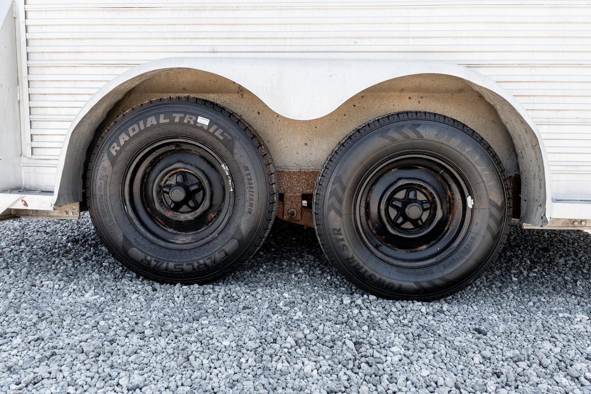 Two black trailer wheels with rims under a white corrugated trailer, on a gravel surface, showing rust and dirt around the axles.