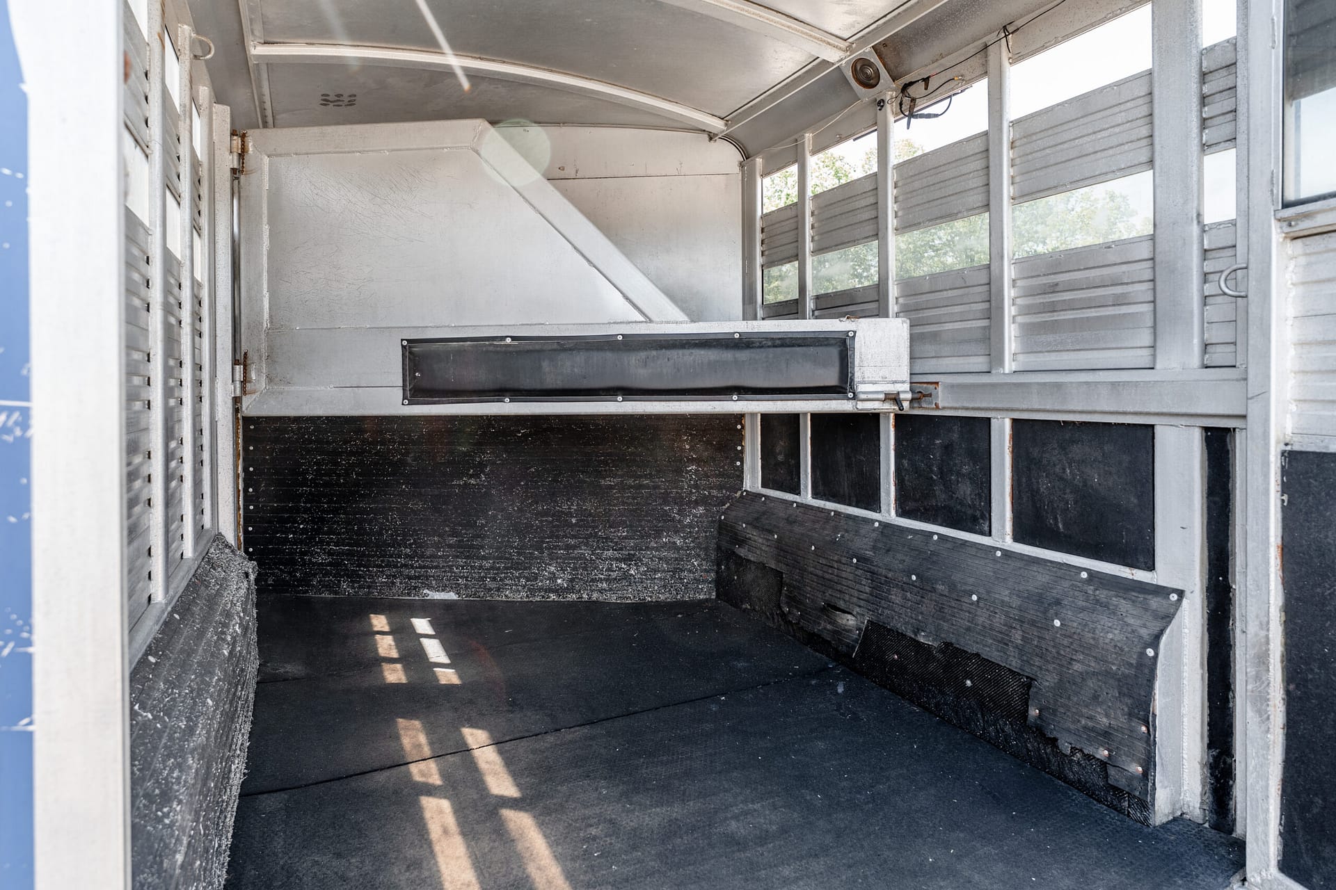 Interior of an empty metal trailer with slatted side panels, a diagonal support beam, and a black rubber floor mat. Light filters in from outside.
