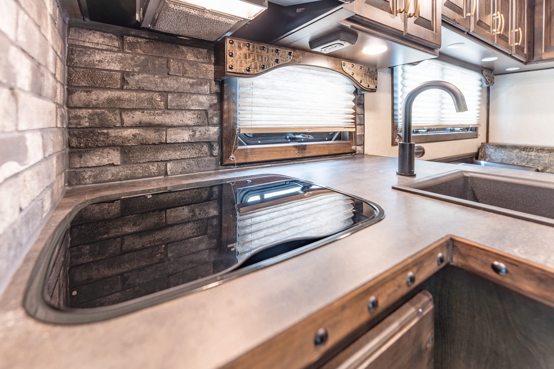 RV kitchen interior featuring a stainless sink and curved black faucet, brown wood cabinets, and a brick-style wall backdrop with window blinds above