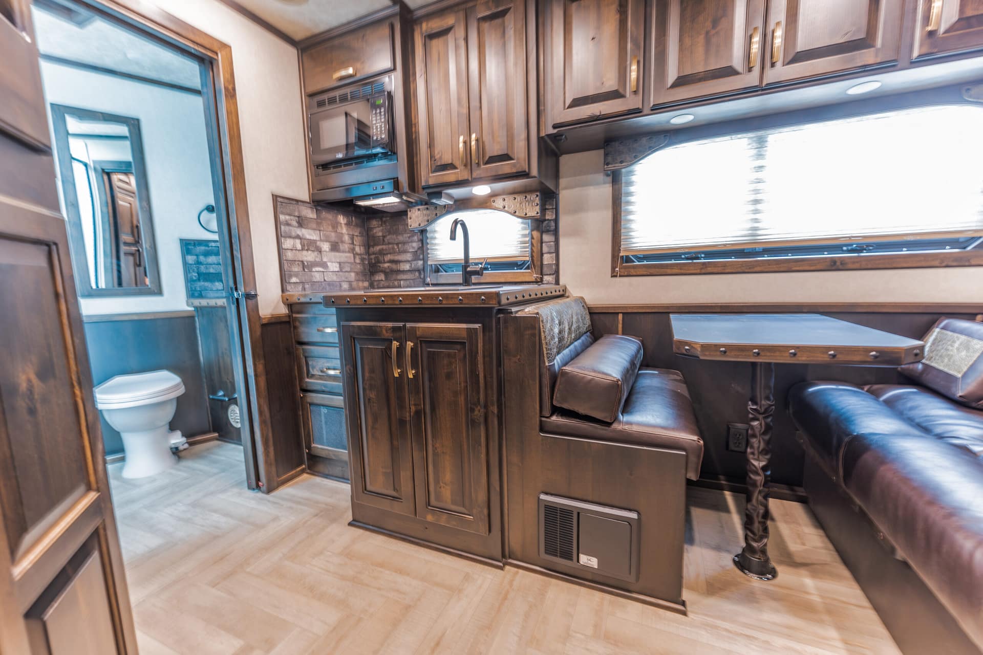 Interior of a luxury RV kitchen with dark wood cabinets, brick backsplash, and built-in microwave above the counter; a sink under the window and brown leather seating nearby.