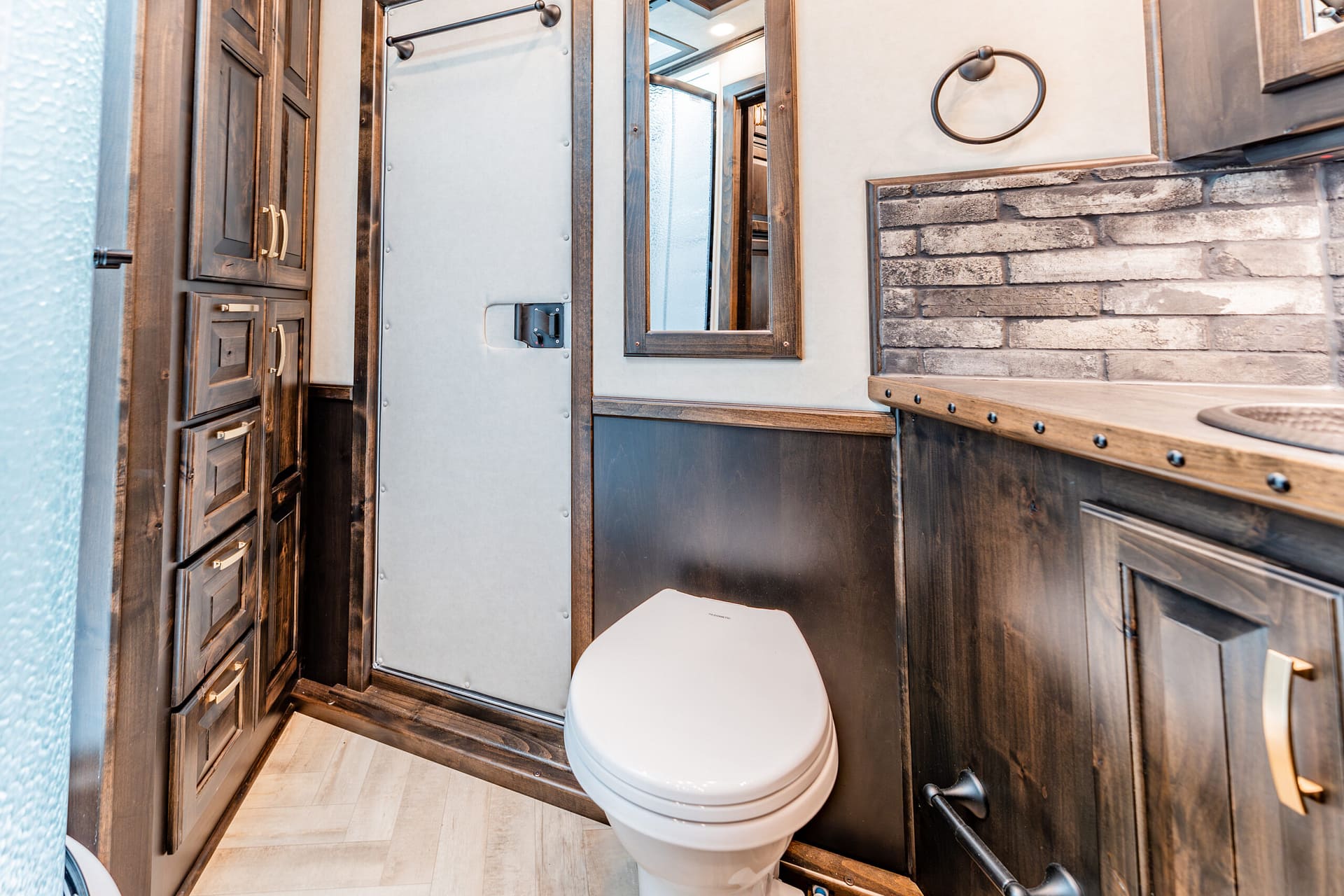 White toilet in a compact rustic bathroom with dark wood cabinets and brick accents on the wall, round mirror above the counter, and a towel ring nearby
