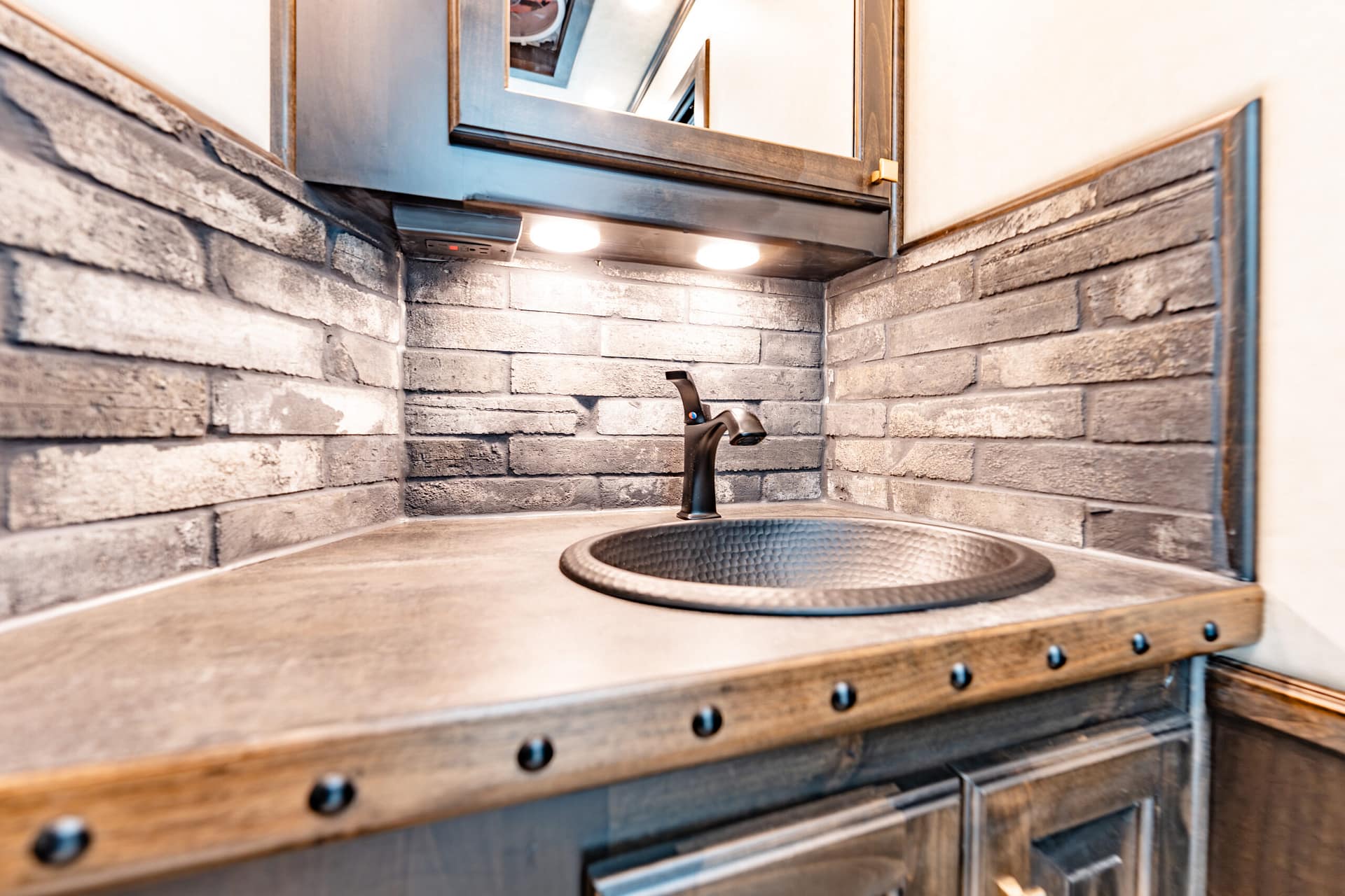 Outdoor grill area with brick backsplash, circular sink, black faucet, and under-cabinet lights above a metal countertop.