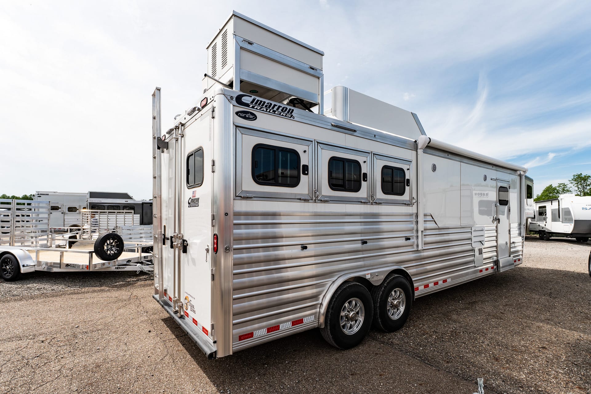 Silver horse trailer with multiple side windows and chrome trim, parked in a lot with other trailers visible in the background (Cimarron Trailers logo).