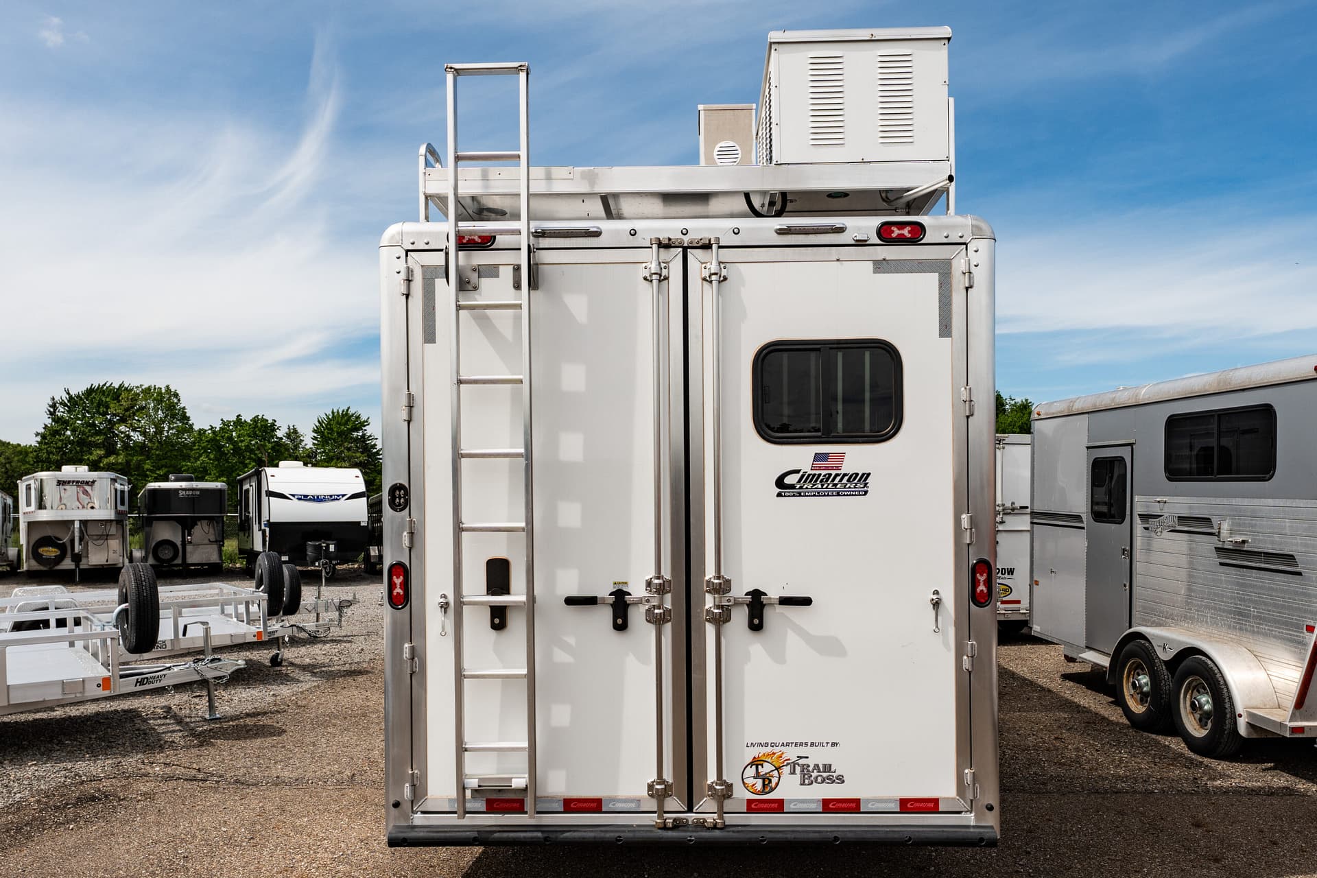 Back view of a white cargo trailer with a vertical ladder, dual doors, and a small flag sticker, parked in a trailer yard under a blue sky.