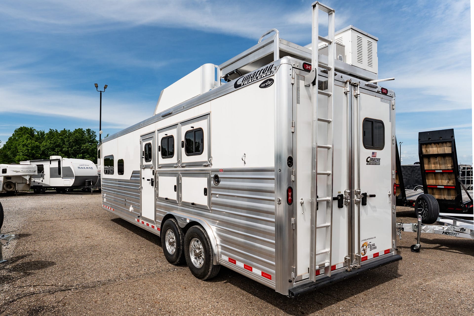 Large silver aluminum trailer with multiple side doors and a rear ramp in a parking lot.