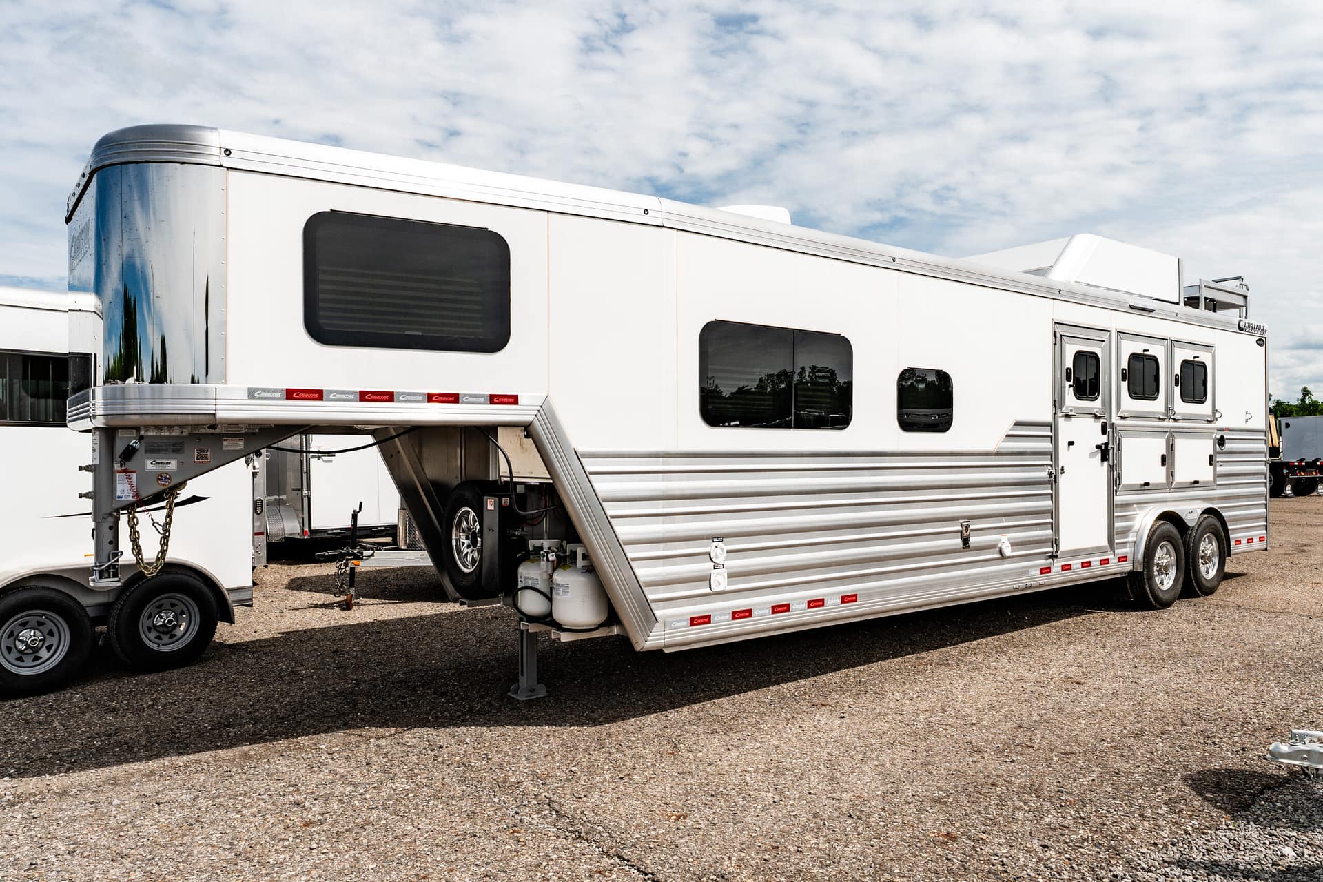 Large white horse trailer with windows and a raised entry stair on a gravel lot; propane tanks nearby.