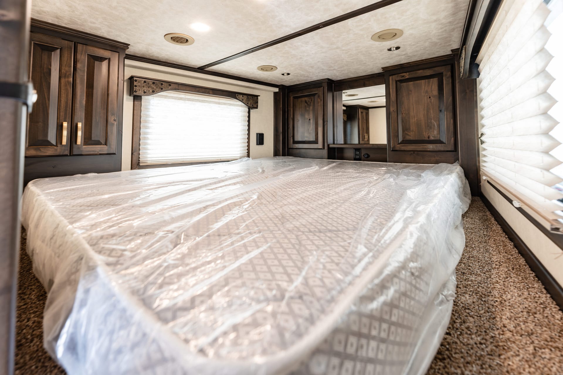 RV bedroom with dark wood cabinets, a window with blinds, and a bed covered in plastic