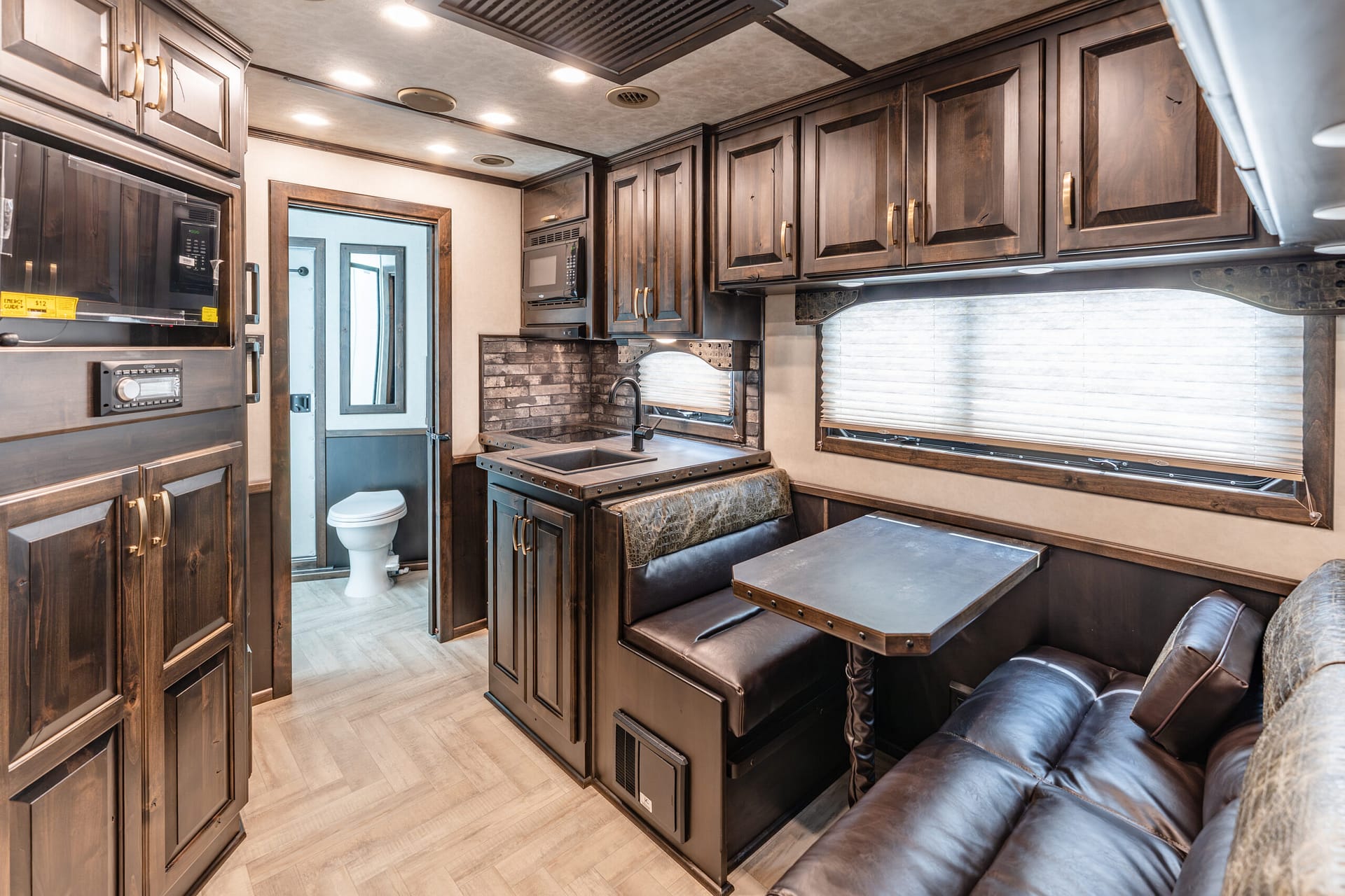 Dark wood RV kitchen with built‑in appliances (microwave, oven) and a cozy brown leather dining area; large window with blinds on the right.