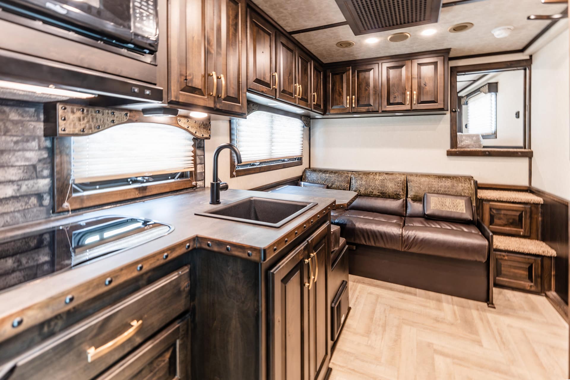 RV kitchen with dark wood cabinets, stainless sink, black faucet, and a sofa area beneath window blinds in a travel trailer.