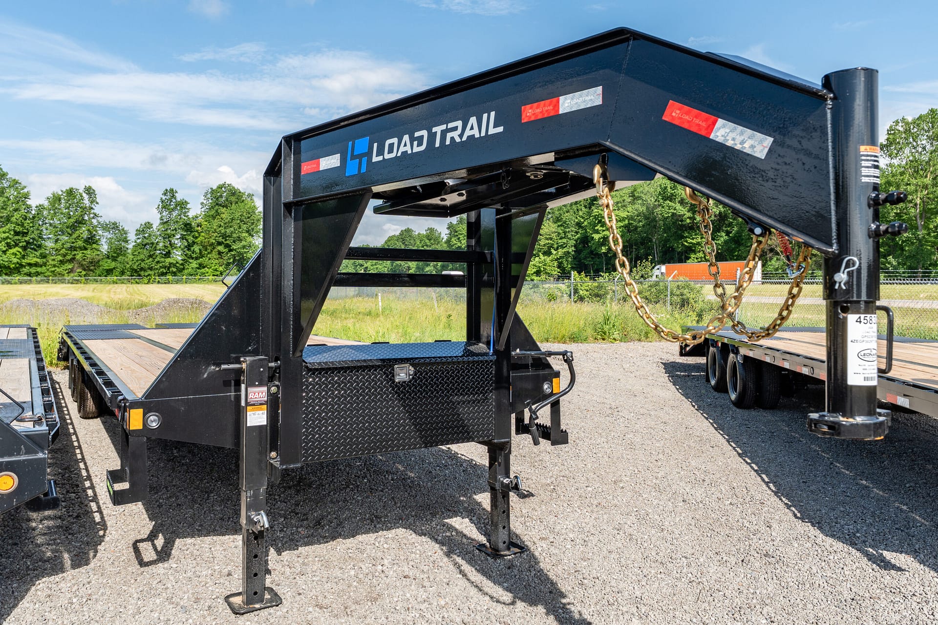 Black Load Trail trailer frame on a gravel lot, with hanging chains and support legs, blue sky and trees in the background.