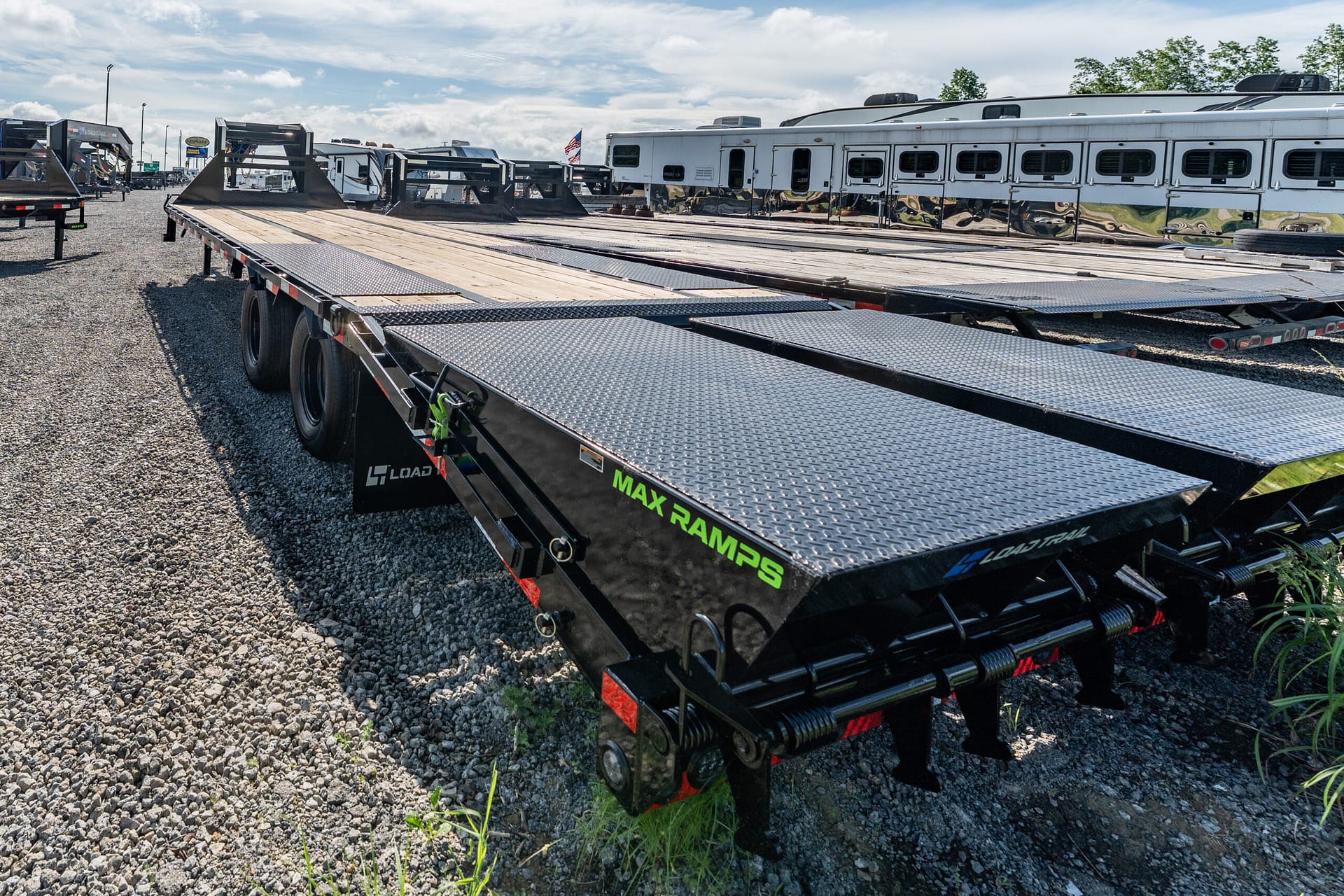 Long black flatbed trailer with diamond-plate ramps in a gravel lot, with other trailers and a silver horse trailer in the background.