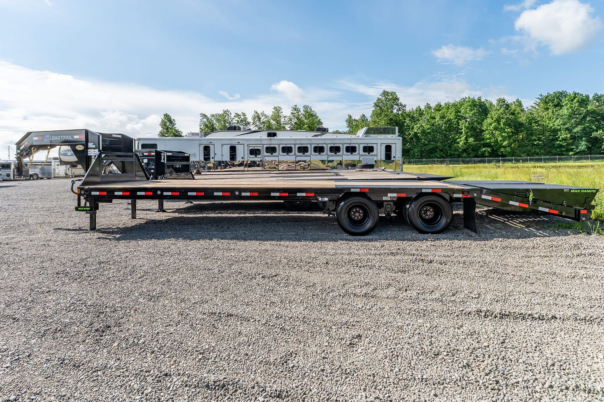 Long flatbed trailer on a gravel lot with a white horse trailer and trees in the background under a blue sky.