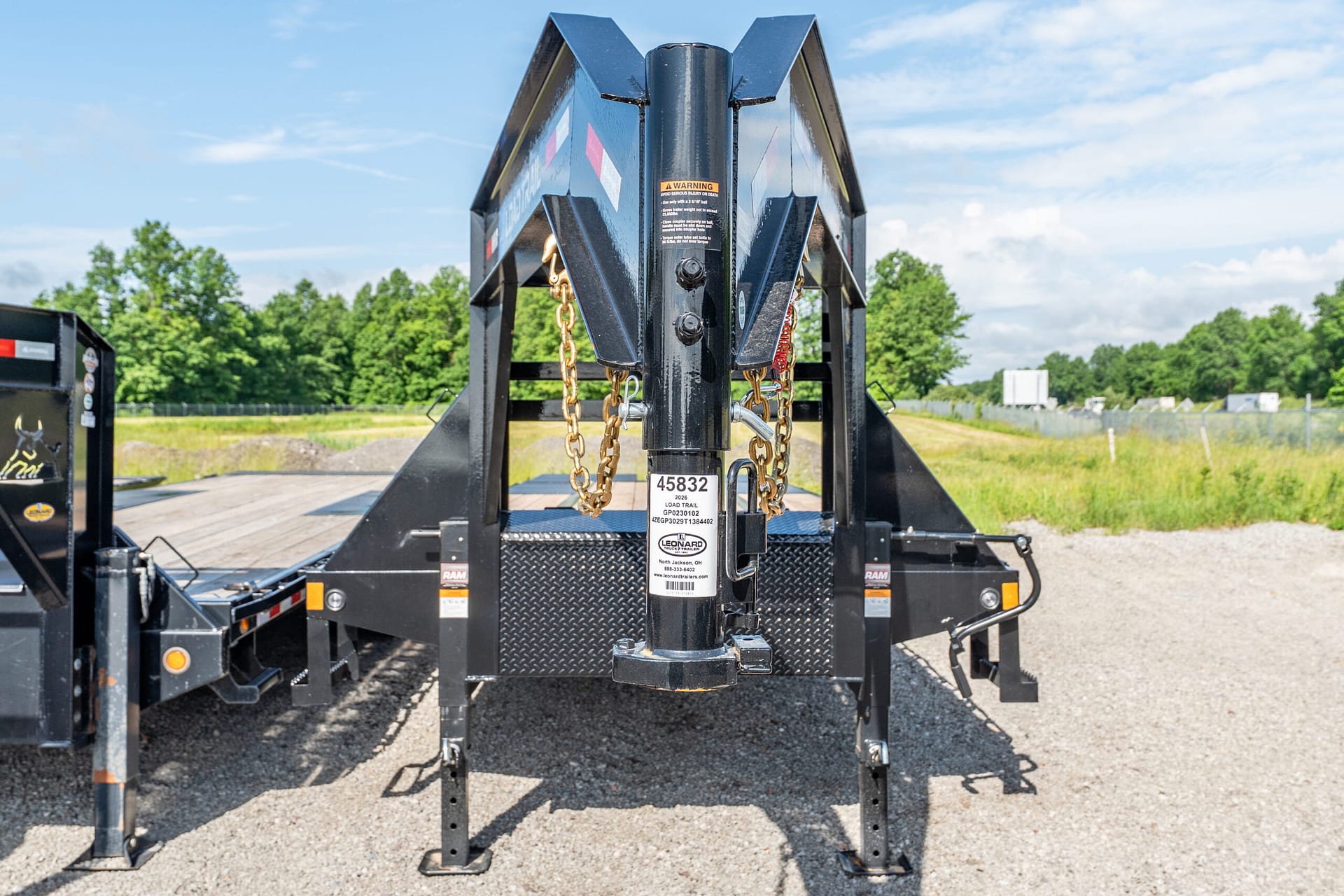 Close-up of a heavy-duty trailer hitch with gold safety chains and a central vertical post, on a gravel lot with blue sky and trees in the background
