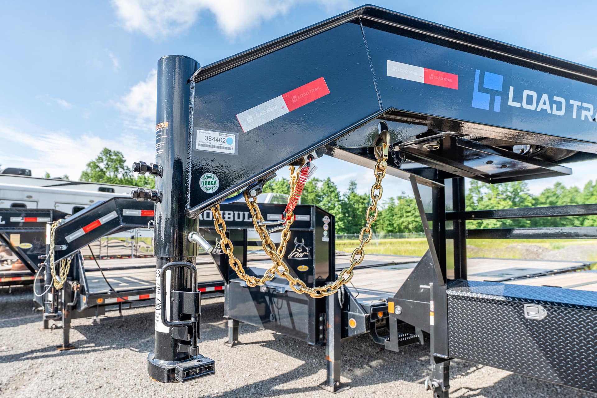 Close-up of a black trailer hitch with gold safety chains and a pin, Load Trail branding visible, in a gravel lot with other trailers in the background.