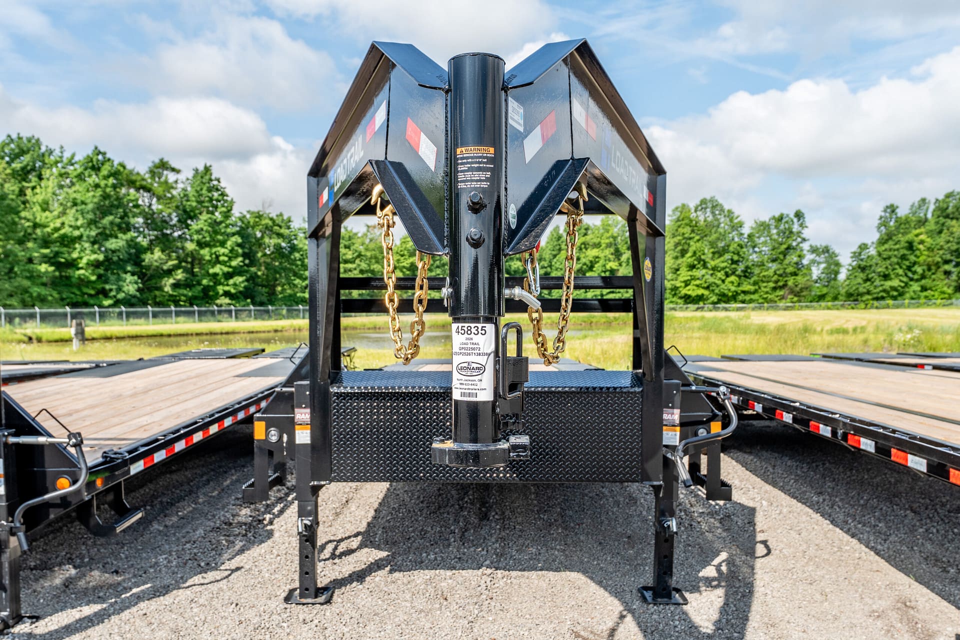 Close-up of a heavy-duty trailer hitch with safety chains on a flatbed trailer, metal frame and wood decking, outdoors with trees and a blue sky in the background