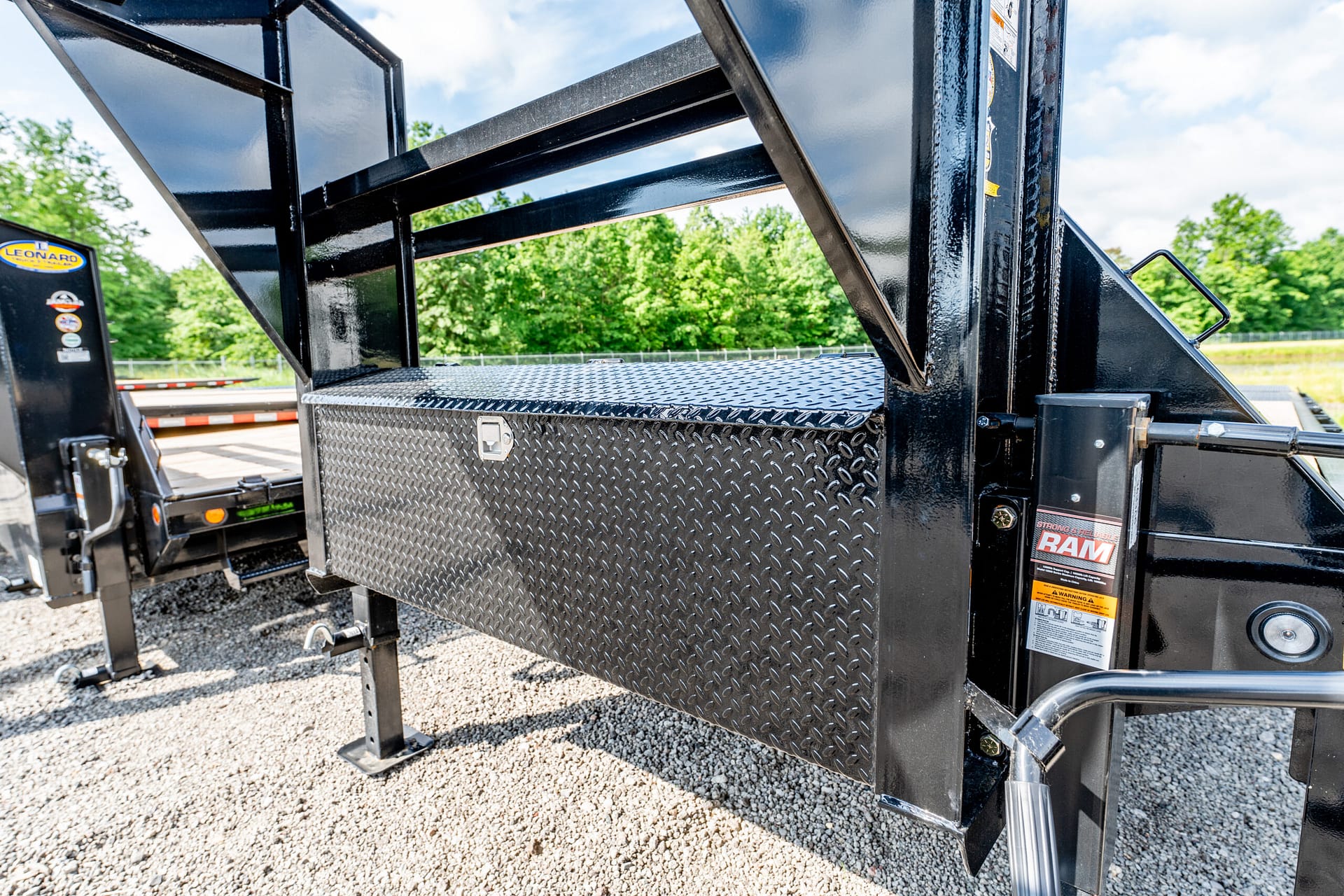 Close-up of a black metal trailer with a diamond-plate toolbox, RAM branding, and hydraulic components outdoors on a gravel lot.