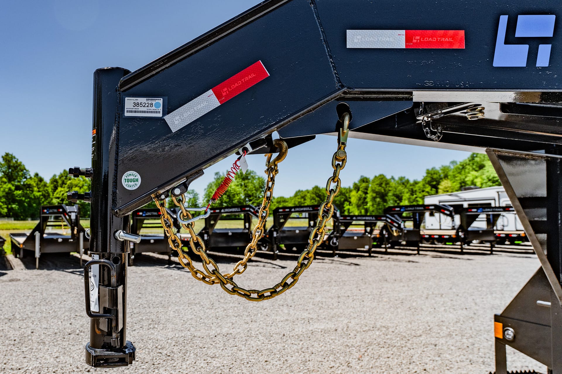 Close-up of a black trailer hitch with gold safety chains and a red Load Trail tag, against a gravel lot.