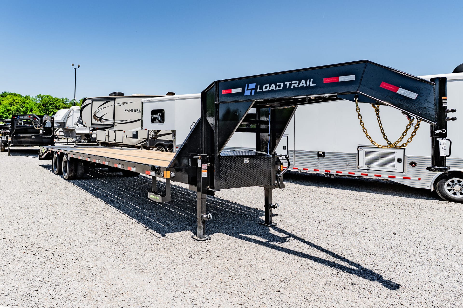 Flatbed trailer being loaded in a gravel lot with a blue sky; visible Load Trail gooseneck trailer and white RVs in the background.
