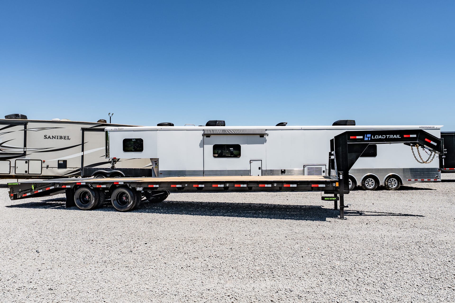 Row of large white recreational trailers on a gravel lot under a clear blue sky; branding visible on the sides.