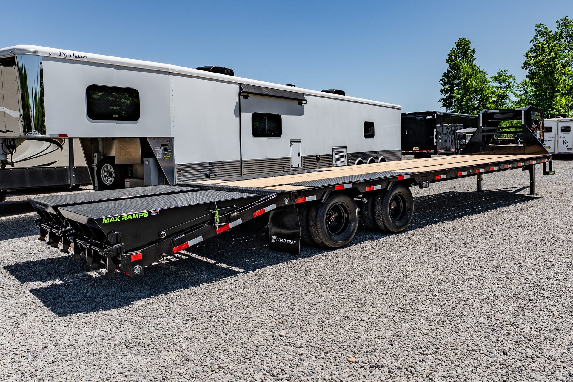 White toy hauler RV mounted on a flatbed trailer in a gravel lot with trees in the background.”