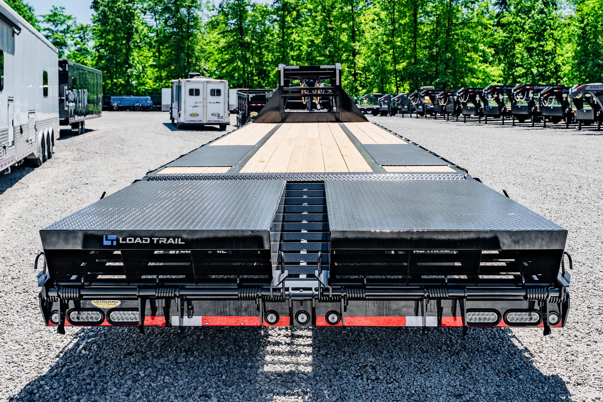 Rear view of a Load Trail car hauler trailer in a gravel lot with other trailers and a green, tree-lined background at the far edge of the yard.