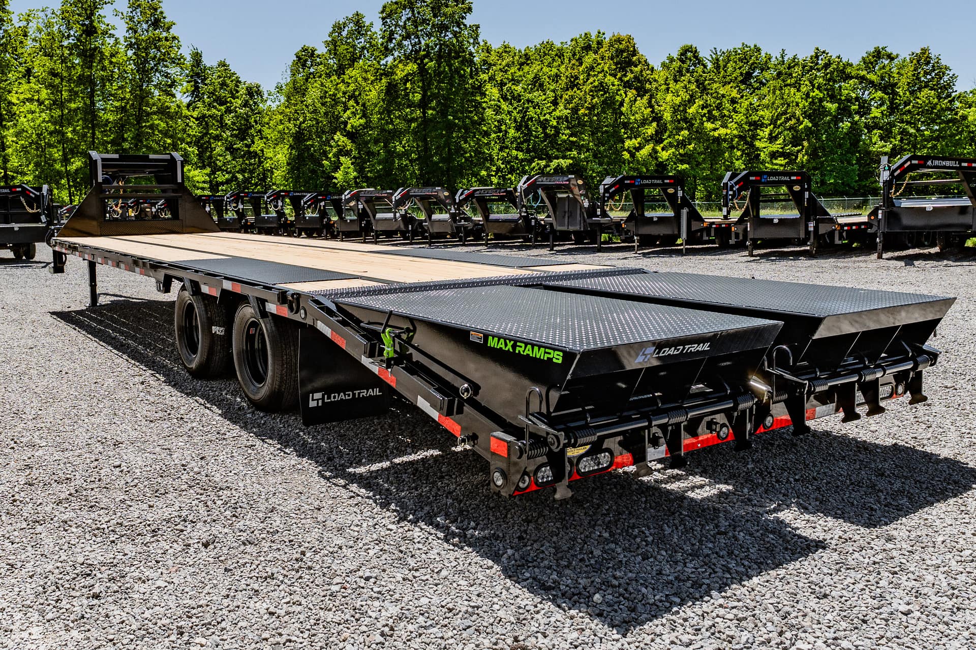 Long black Load Trail flatbed trailer with multiple loading ramps, parked on a gravel lot with green trees in the background.
