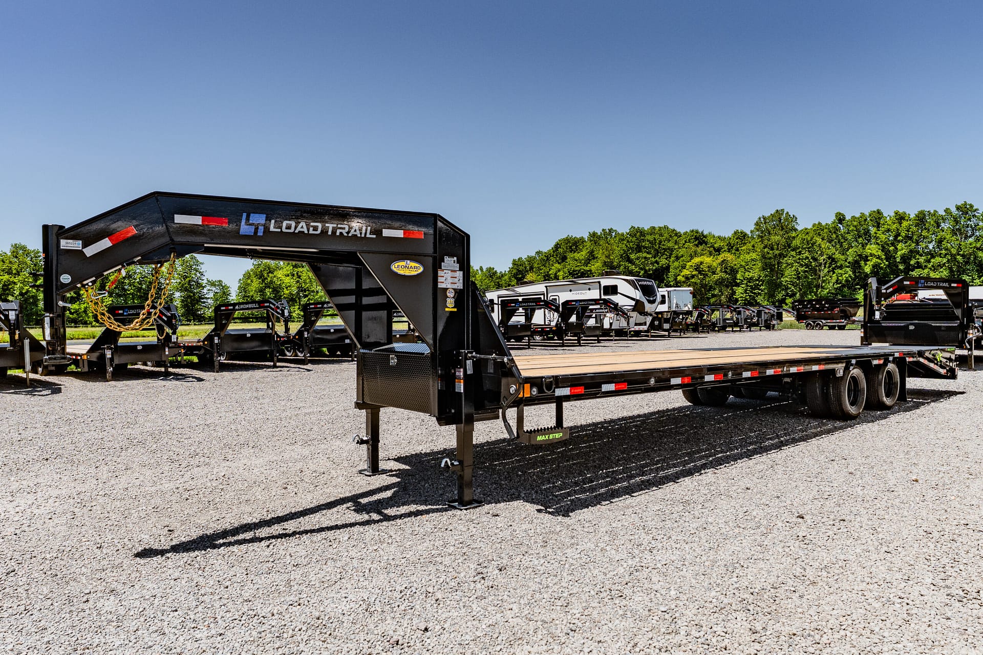 Wide view of a trailer lot with a black Load Trail gooseneck trailer in the foreground and rows of trailers and a white RV in the background under a clear blue sky.