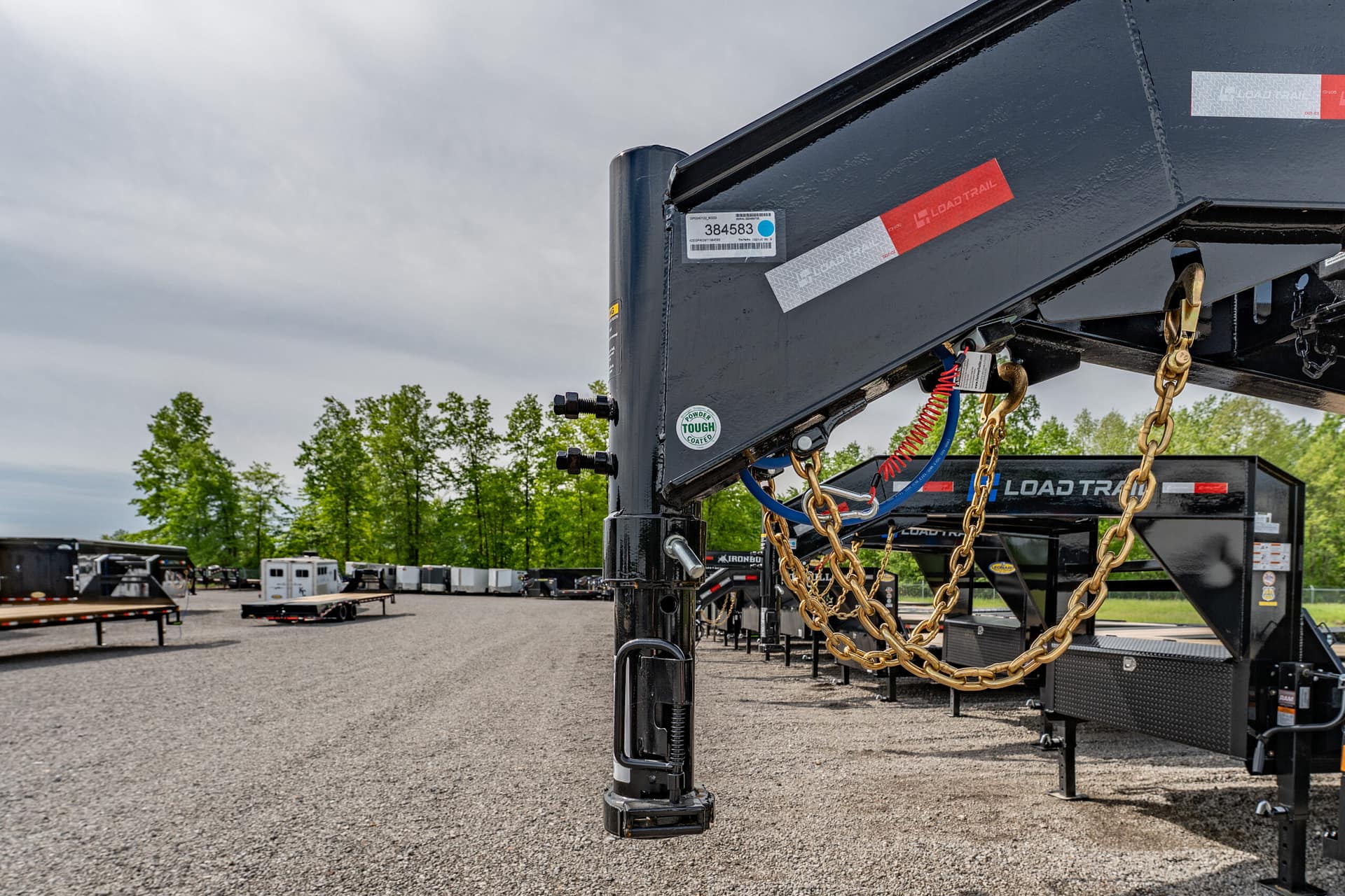 Close-up of a black trailer hitch with gold chains and safety pins, in a lot of trailers and equipment against a cloudy sky backdrop.