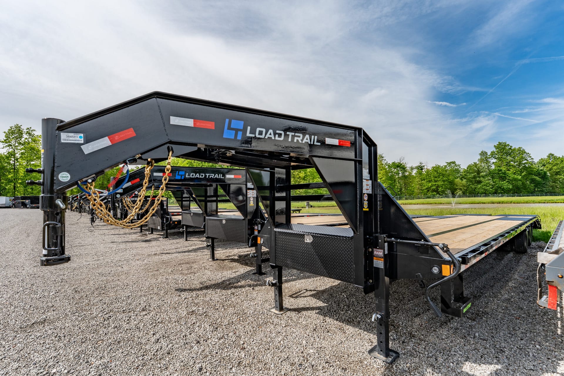 Black Load Trail trailer frame with wooden deck and hanging safety chains on a gravel lot under a blue sky with clouds, outdoor equipment yard behind it.