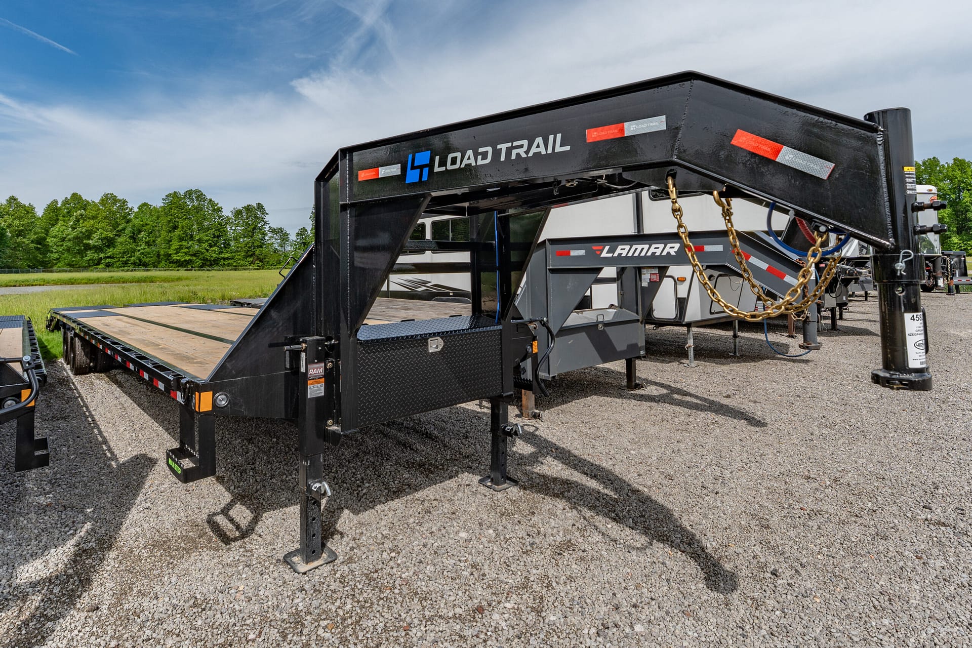 Black Load Trail trailer frame with a flatbed deck and safety chains on a gravel lot under a blue sky.