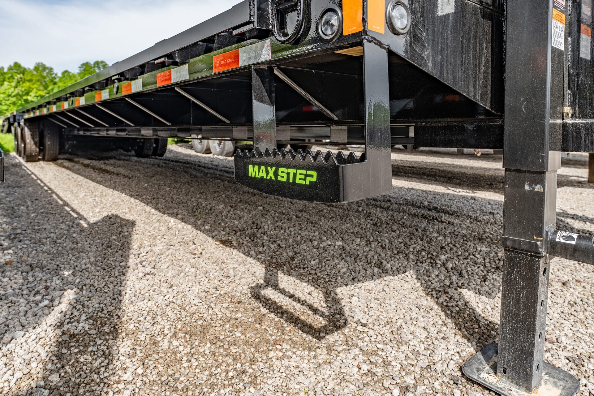 Underside of a black trailer showing a protruding MAX STEP stair on a gravel surface under the trailer’s frame.