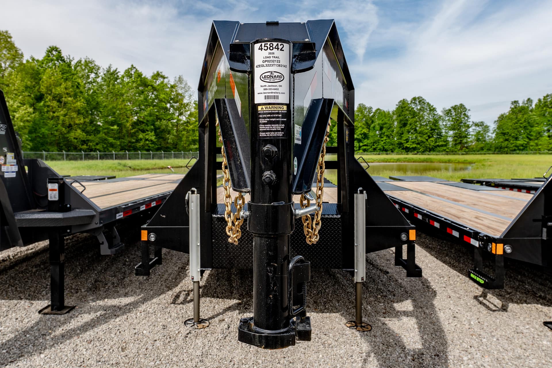 Close-up of a heavy trailer hitch with chains, labels, and a black tow bar against gravel ground and green trees in the background.