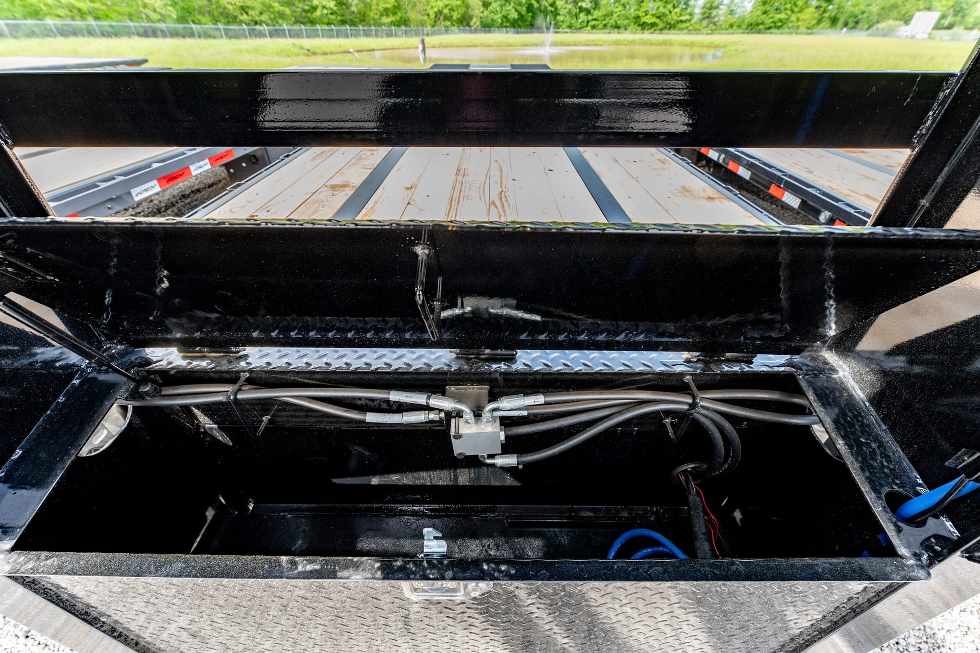 View from inside a black trailer frame showing wooden deck planks, exposed hydraulic lines, and metal supports; outdoors with grass and fence in distance.