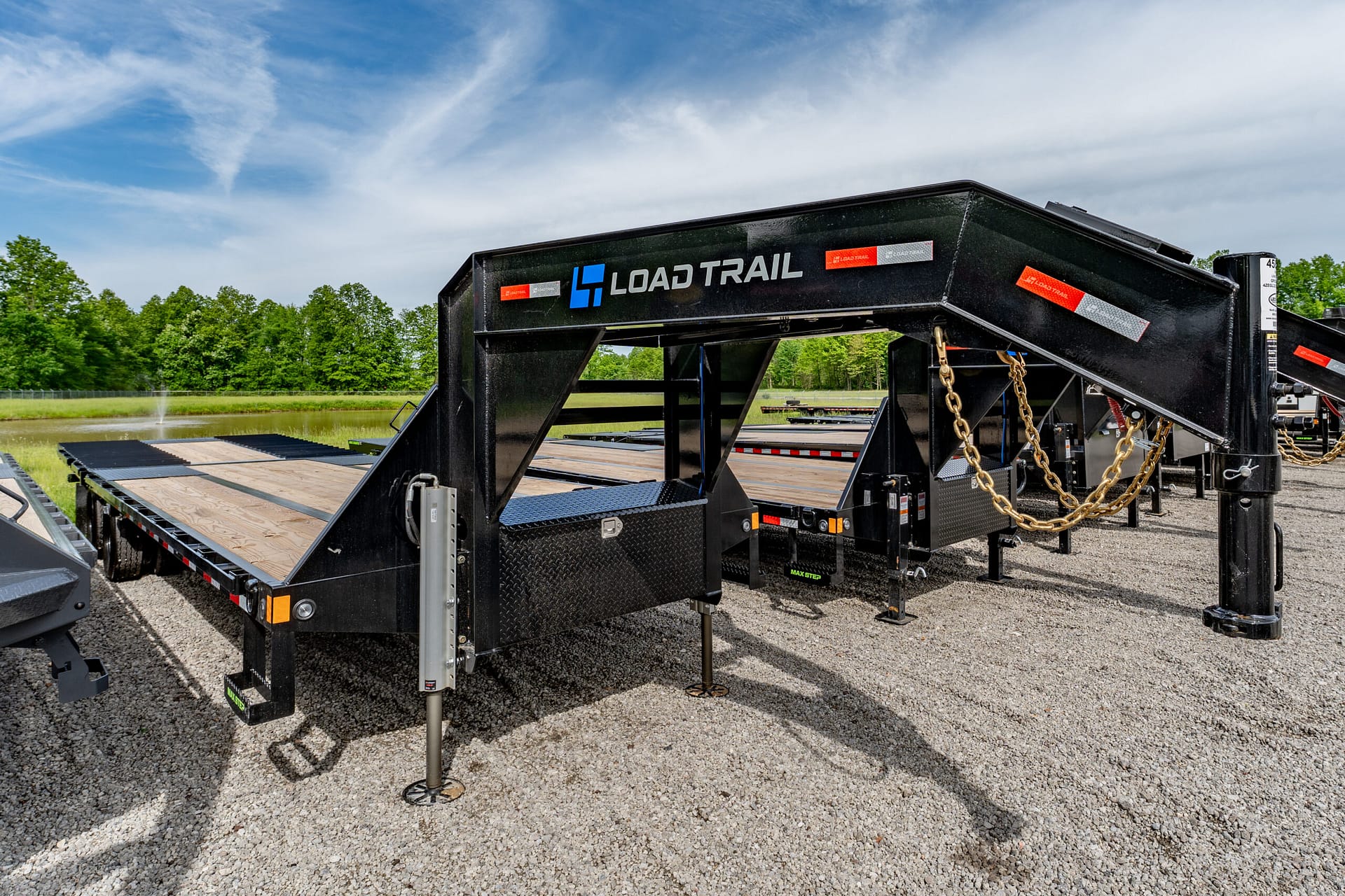 Black Load Trail gooseneck trailer with a wooden flatbed in a gravel lot under a blue sky, trees in the background.