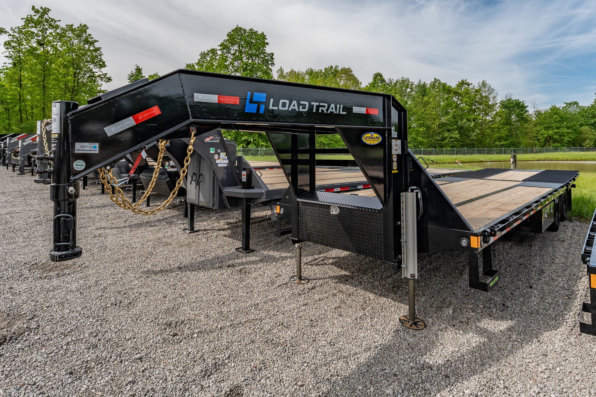 Black Load Trail trailer with wooden deck and safety chains on a gravel lot, trees and blue sky in the background.”