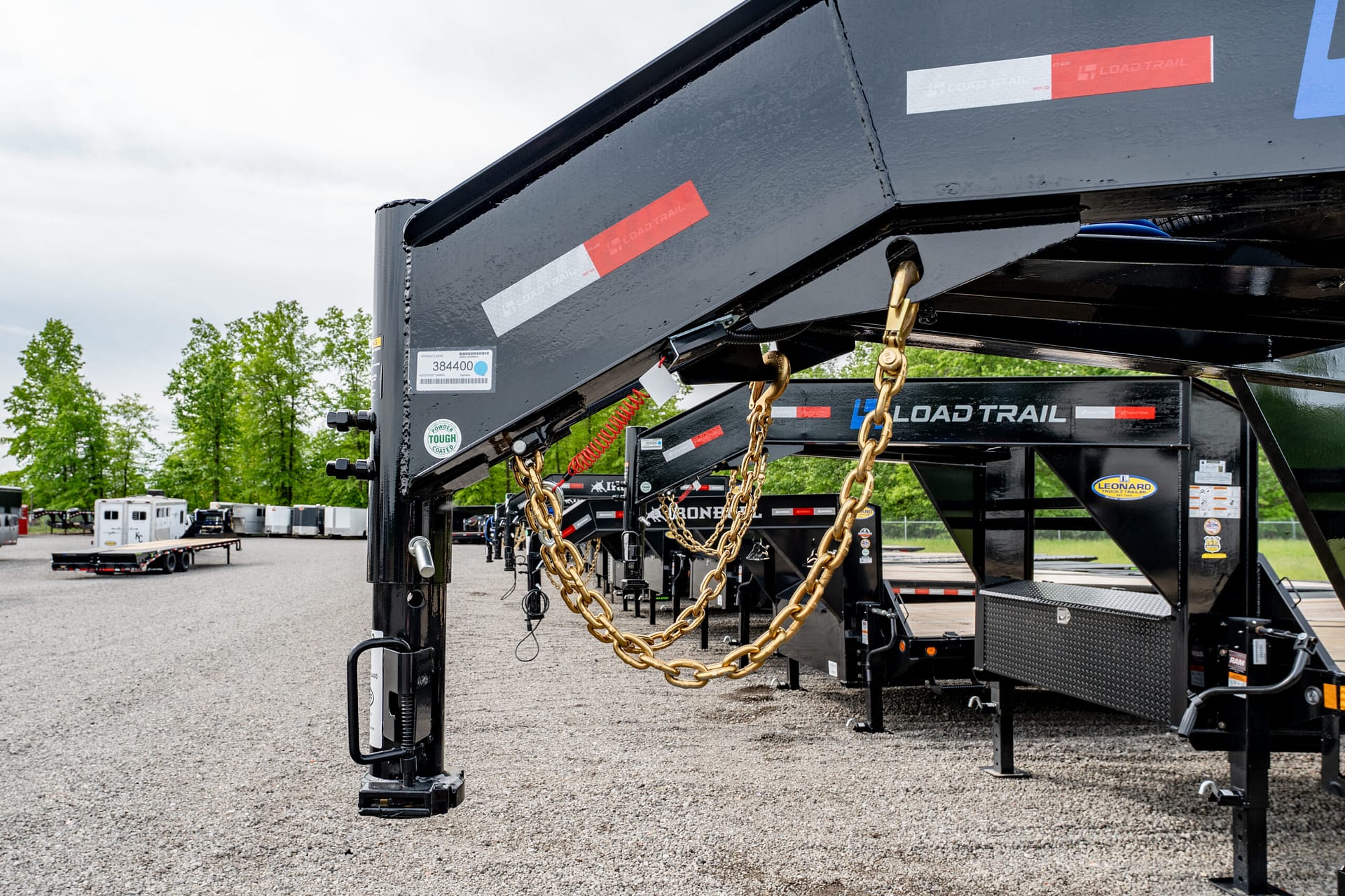 Close-up of a black Load Trail trailer frame with heavy gold chains hanging from the hitch in a gravel lot.