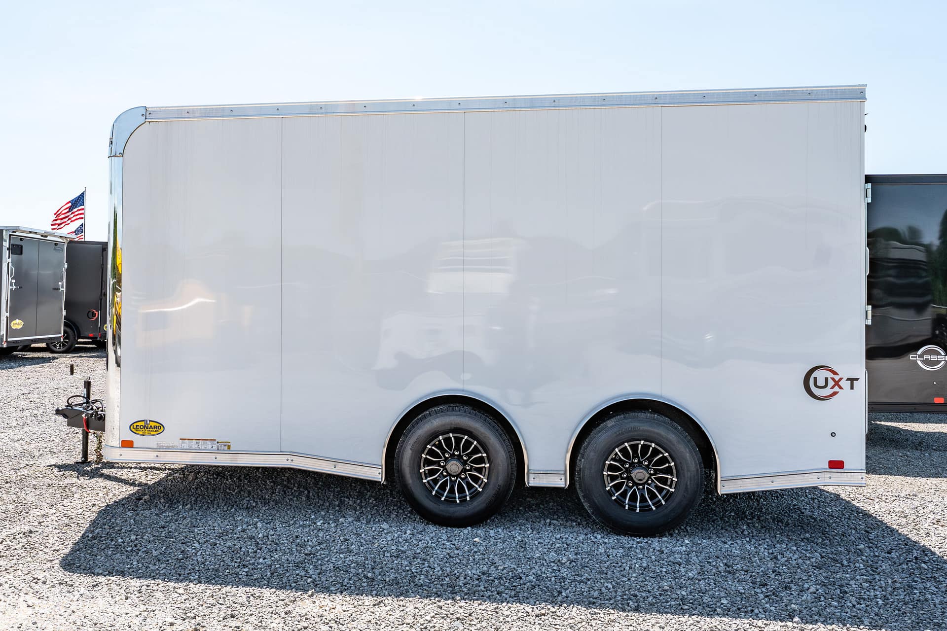 Side view of a large silver enclosed trailer with triple axle wheels, parked on a gravel lot; a small American flag is visible in the background.