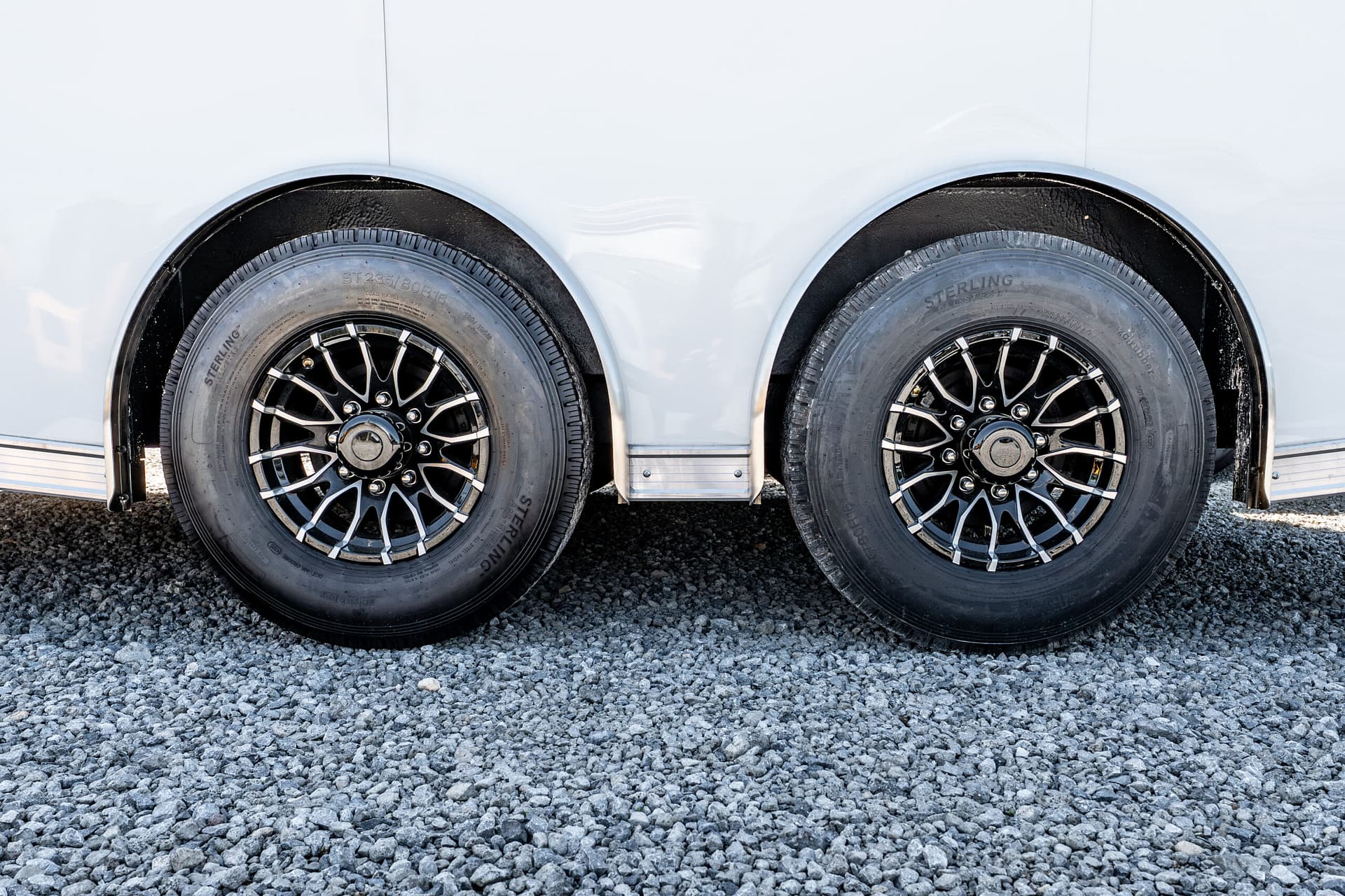 Pair of trailer wheels with black alloy rims under a white trailer, resting on gravel ground.