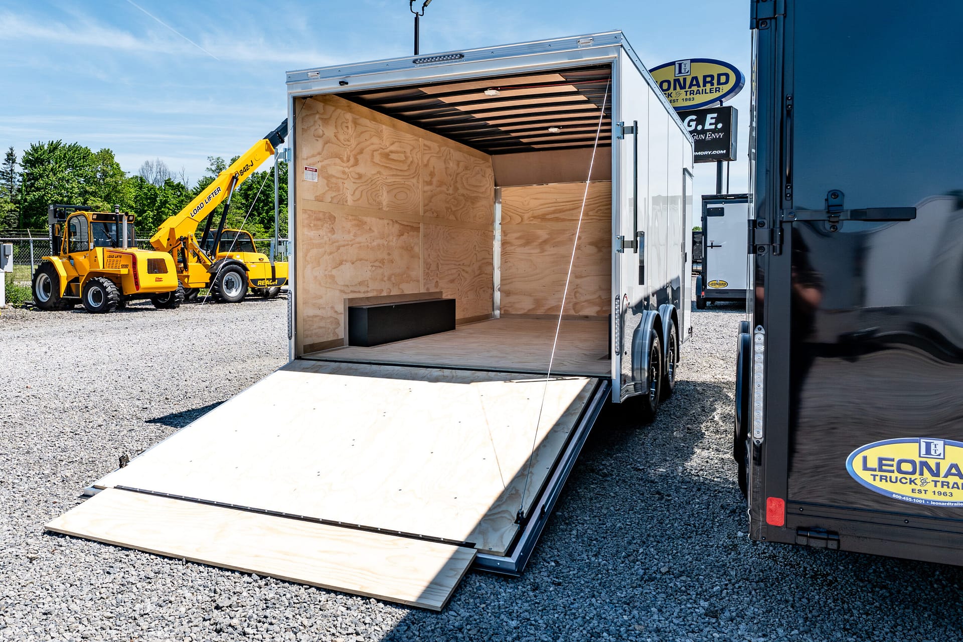 Open loading trailer with plywood walls and a lowered wooden ramp, flanked by yellow construction equipment on a gravel lot.